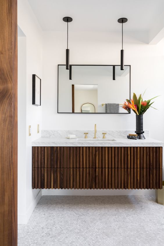 A dark stained fluted vanity with a stone countertop, a mirror in a black frame, black pendant lamps and bold blooms