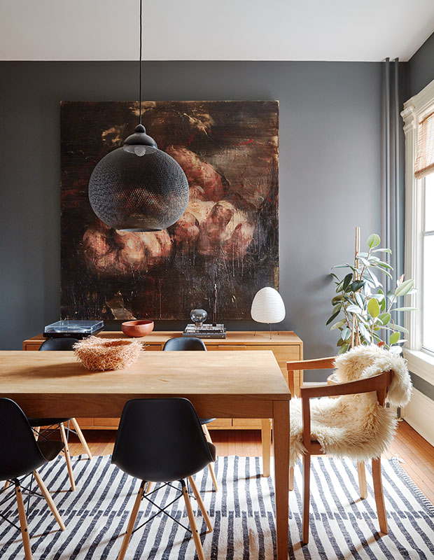 A dining room with soot walls, light stained furniture, soot chairs, a vintage artwork and a pendant lamp
