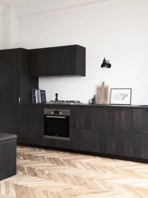 A dramatic dark stained sleek kitchen with no handles and built in appliances plus some decor, a black sconce on the wall