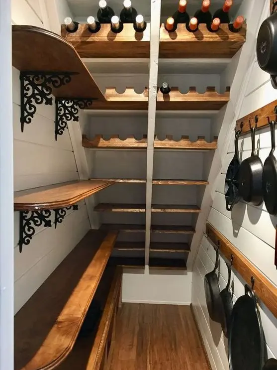 A farmhouse stairs pantry with open shelves with wrought details, wooden rails with hooks and built in stained shelves and wine bottle stands