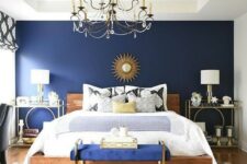a glam bedroom with a navy wall, a wooden bed, a bold blue bench, gold nightstands and a tray plus a vintage chandelier