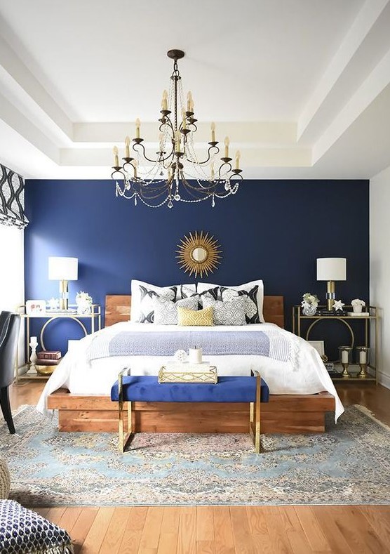 a glam bedroom with a navy wall, a wooden bed, a bold blue bench, gold nightstands and a tray plus a vintage chandelier