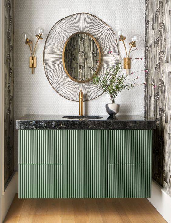 a green fluted vanity with a black stone countertop, a mirror in a unique frame, creative flower-like sconces and greenery