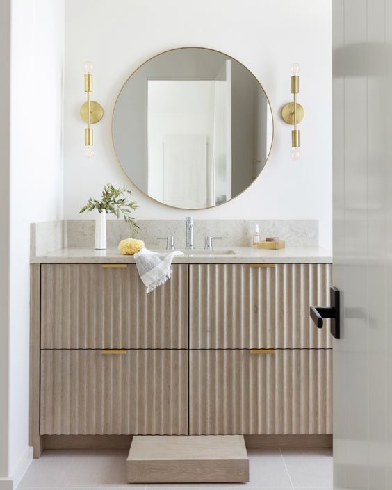 a greige fluted vanity with gold handles, a stone countertop, a round mirror, gold sconces and some lovely decor