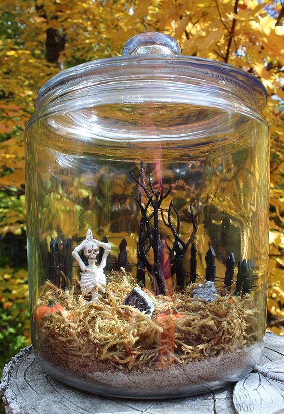 a jar with greenery, mini pumpkins, tombstones and a skeleton is a cool mini terrarium for Halloween, you can repeat one yourself