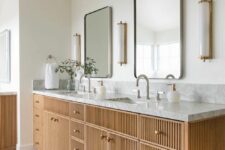 a large fluted double vanity with a stone countertop, brass knobs, stylish mirrors, sconces and greenery