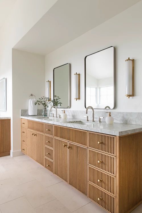 a large fluted double vanity with a stone countertop, brass knobs, stylish mirrors, sconces and greenery