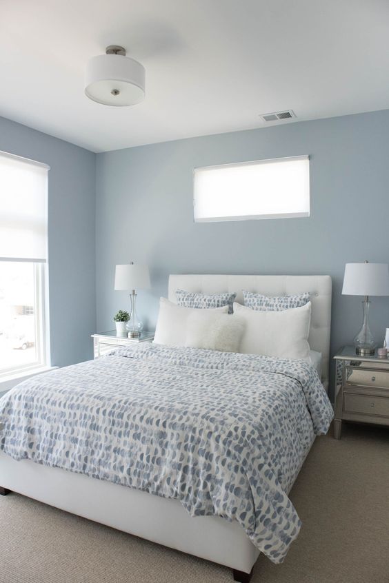 a light blue bedroom with windows, a white bed with blue and white bedding, mirror nightstands and table lamps