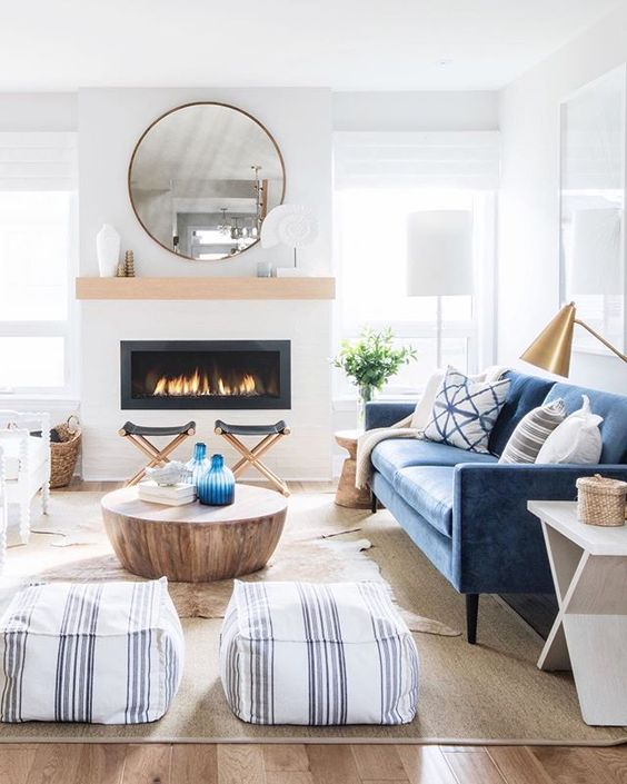 A light filled coastal living room with a built in fireplace, a blue sofa, a coffee table, printed poufs, folding stools and a gold lamp
