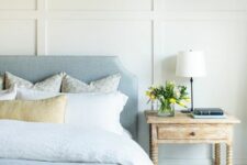 a lovely coastal bedroom with paneled walls, a pastel blue bed and neutral bedding, a blue ottoman and a shabby chic nightstand