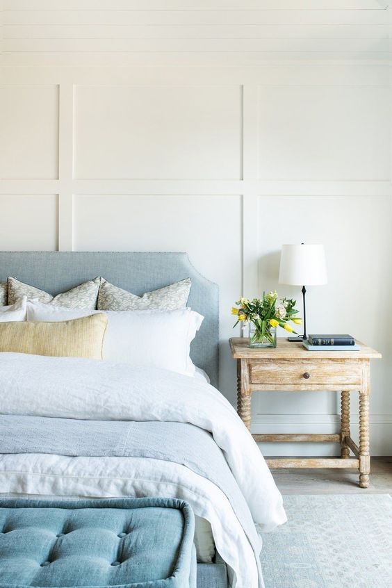 a lovely coastal bedroom with paneled walls, a pastel blue bed and neutral bedding, a blue ottoman and a shabby chic nightstand