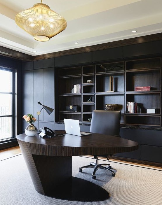 A minimalist and refined home office with a black built in shelving unit, a sculptural oval desk of rich stained wood, a bold chandelier