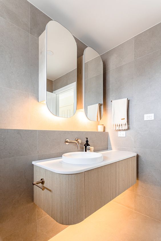 a minimalist bathroom clad with stone tiles, with a curved fluted vanity with lights, a vessel sink, rounded mirrors with lights