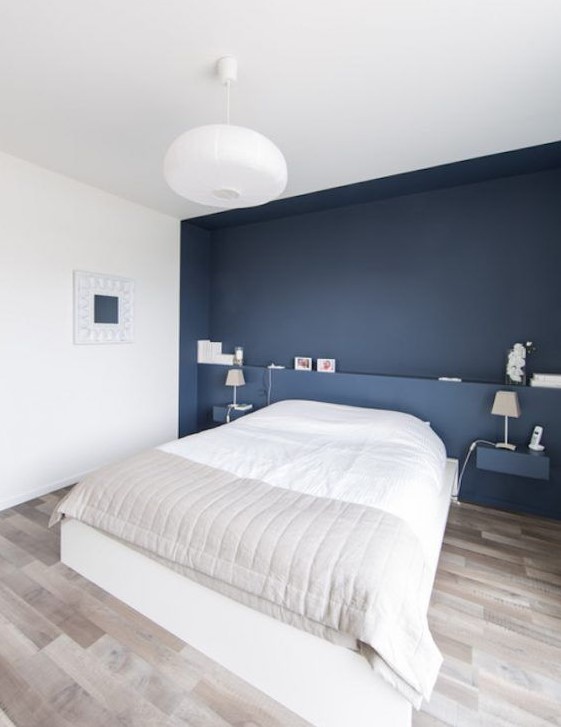 a minimalist bedroom with a navy accent wall and floating nightstands, a white bed and a pendant lamp plus an artwork