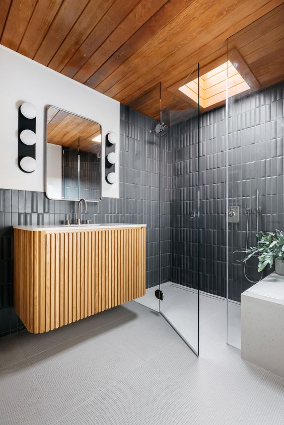 a modern bathroom with graphite grey skinny tiles, a shower space with a skylight, a fluted floating vanity and a mirror with lights