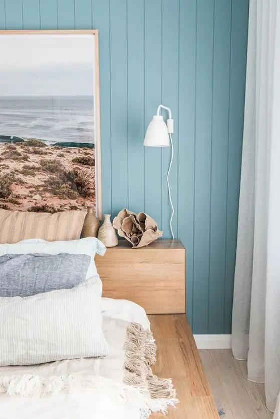 a modern beach bedroom with blue walls, a wooden bed, a statement artwork and printed and blue bedding