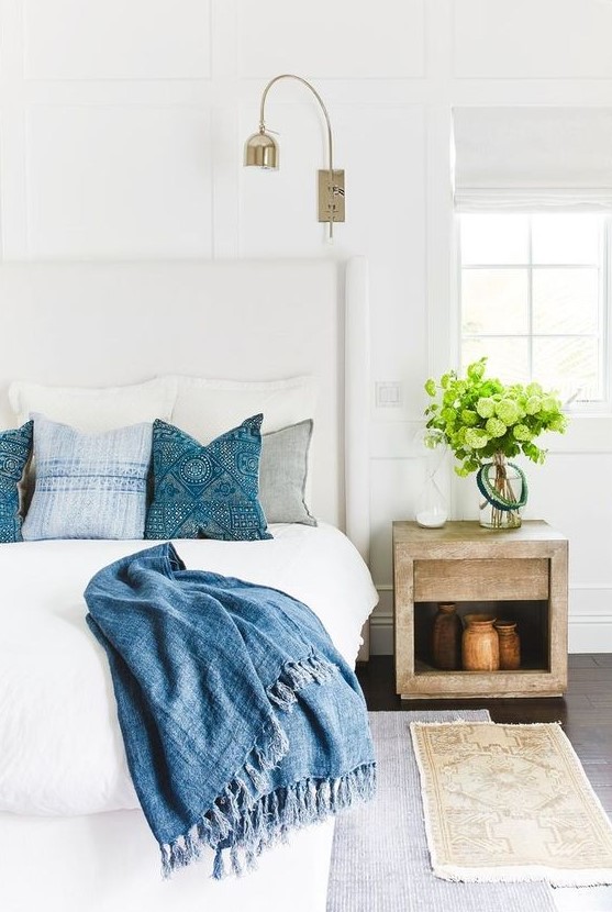 a modern coastal bedroom in neutrals, with a wooden nightstand, layered rugs, greenery and printed blue bedding for a coastal feel