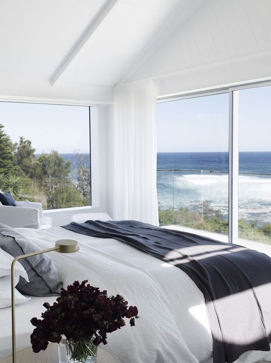 a modern coastal bedroom with glazed walls and a balcony, with white furniture and darker textiles in coastal colors