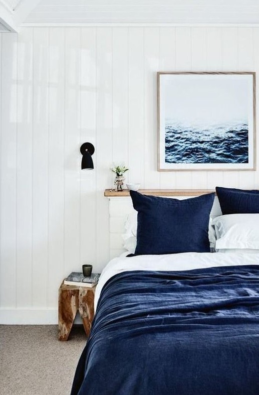 a modern coastal bedroom with navy and white bedding, a sea artwork, black sconces and a wooden stool
