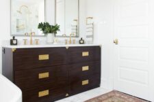 a modern farmhouse bathroom with a double dark-stained vanity with gold fixtures and gold handles, mirrors and gold sconces over them