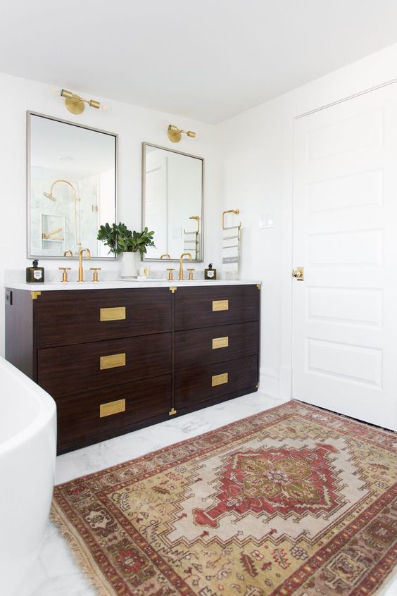 A modern farmhouse bathroom with a double dark stained vanity with gold fixtures and gold handles, mirrors and gold sconces over them
