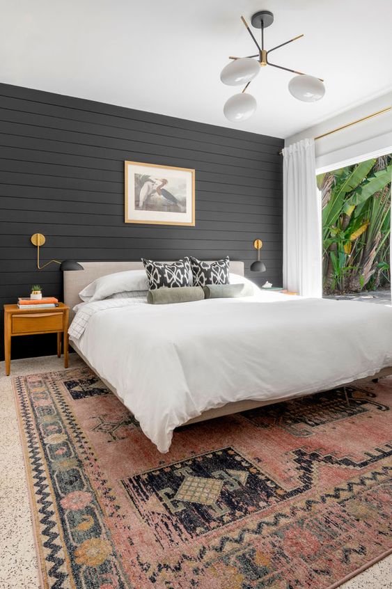 A modern farmhouse bedroom with a black shiplap accent wall, a neutral bed, a printed rug, light stained nightstands and a cool chandelier