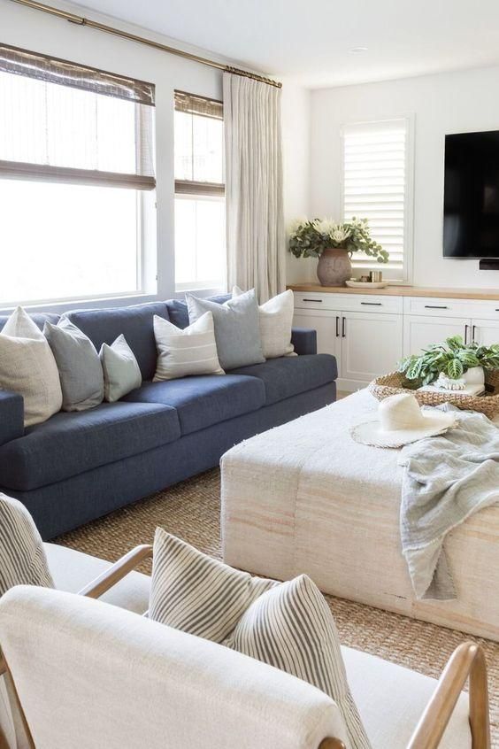A modern farmhouse living room with built in cabinets, a blue sofa, a neutral ottoman, neutral chairs, a jute rug and some greenery
