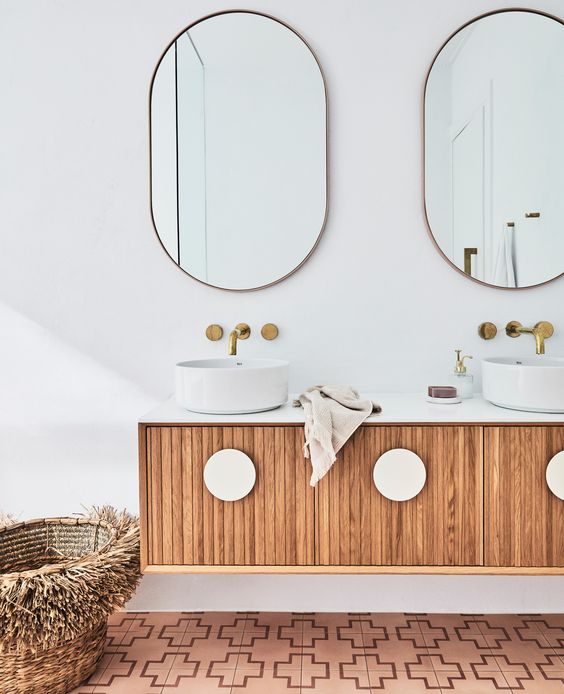 a modern fluted floating vanity with white knobs, oval mirrors, vessel sinks, brass fixtures and a basket