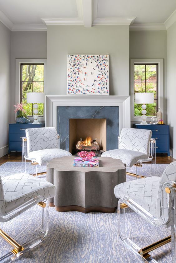 A modern living room with a fireplace clad with marble, blue sideboards, a quirky shaped coffee table, white and acrylic chairs