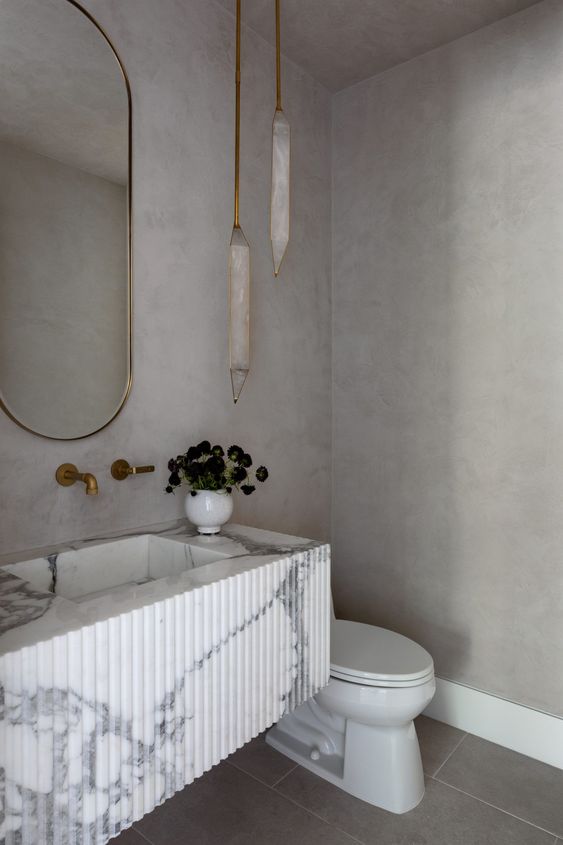 a modern refined powde room done with stone tiles and concrete, with pendant lamps, a reeded vanity with a carved sink and an oval mirror