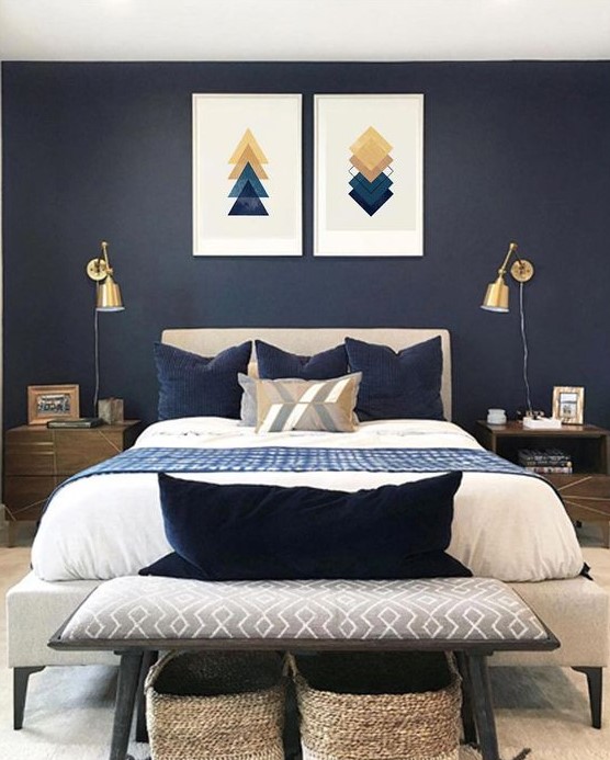 a moody bedroom with navy walls, an upholstered bed, a geometric gallery wall and gold sconces plus a comfy bench