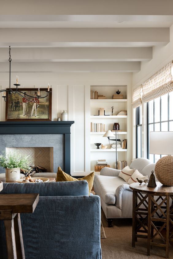 A neutral farmhouse living room with wooden beams, built in shelves, a fireplace with a black mantel, a white sofa, a blue one, a coffee table