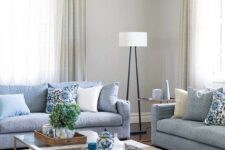 a neutral living room with pale blue sofas, an ottoman, glass coffee tables and printed pillows, a floor lamp