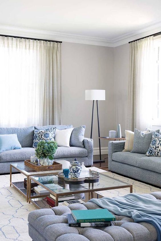 a neutral living room with pale blue sofas, an ottoman, glass coffee tables and printed pillows, a floor lamp