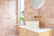 a pink bathroom clad with tiles, with a shower space with a window, a fluted vanity with a terrazzo sink and a round mirror