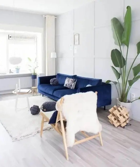 A pretty light filled living room done in white and creamy, with a navy sofa, a couple of side tables, a wooden chair, a potted tree and some wood