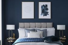 a refined modern bedroom with navy walls, a grey upholstered bed, chic artworks, gold nightstands and blue bedding