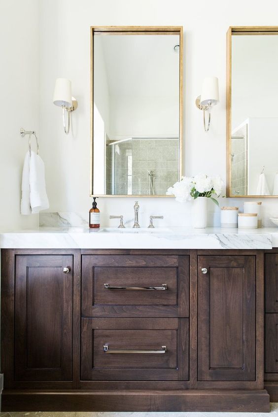 A refined neutral bathroom with a dark stained vanity, a white stone countertop, mirrors in gilded frames and neutral sconces