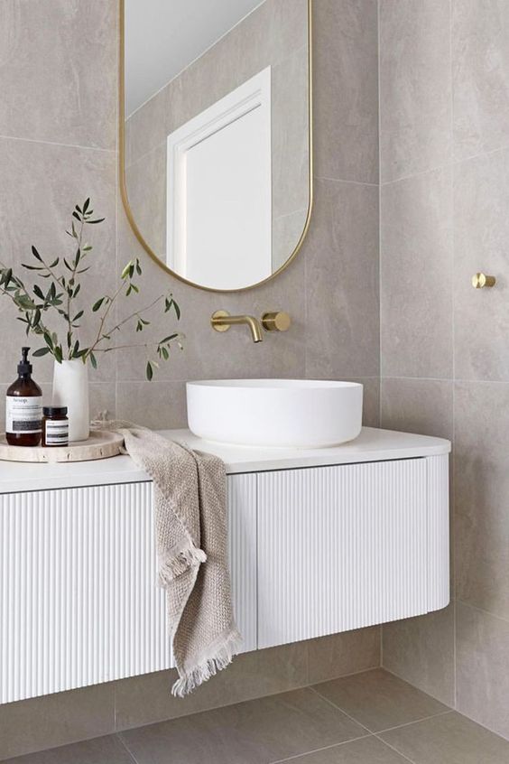 a serene bathroom with grey stone tiles, a white fluted and curved vanity with a vessel sink, an oval mirror and greenery