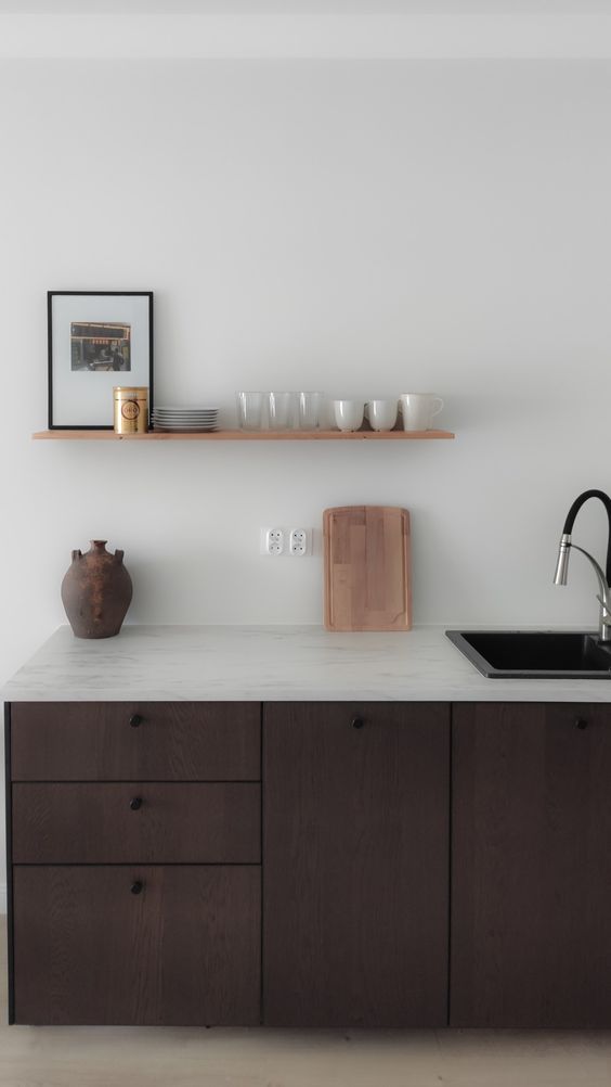 A serene kitchen with dark stained cabinetry, a white stone countertop and a small shelf for holding tableware and dishes
