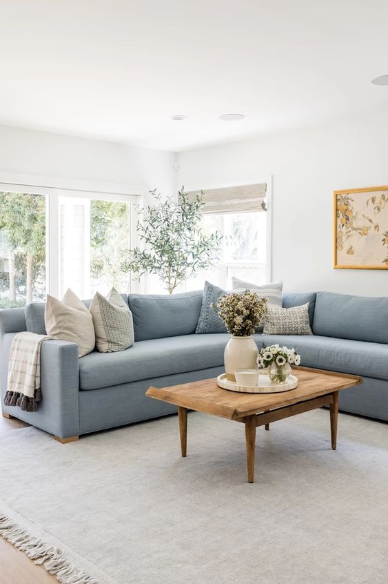 a simple and stylish living room with a glazed wall, a light blue sectional, a wooden coffee table and blooms and greenery