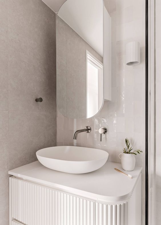 A small and chic nook with a built in white fluted vanity and a vessel sink, an oval mirror cabinet and a vase