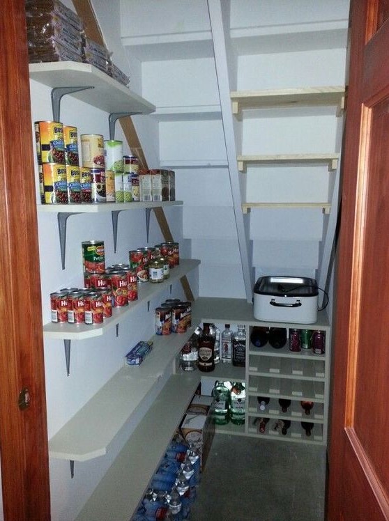 A small and well organized stairs pantry with open shelves and a built in storage unit, with food and drinks stored