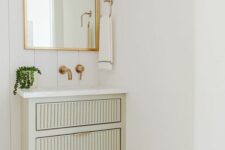 a small pale green fluted floating vanity, an arched mirror in a gold frame, gold fixtures and some greenery for a lovely bathroom