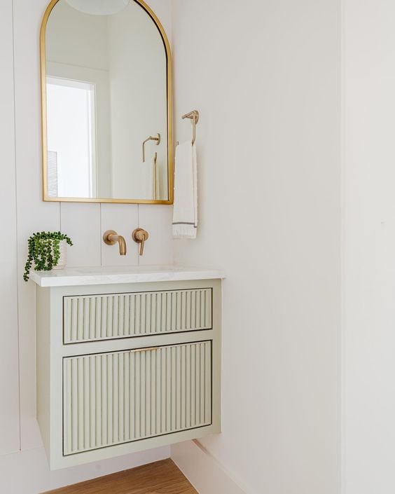 a small pale green fluted floating vanity, an arched mirror in a gold frame, gold fixtures and some greenery for a lovely bathroom