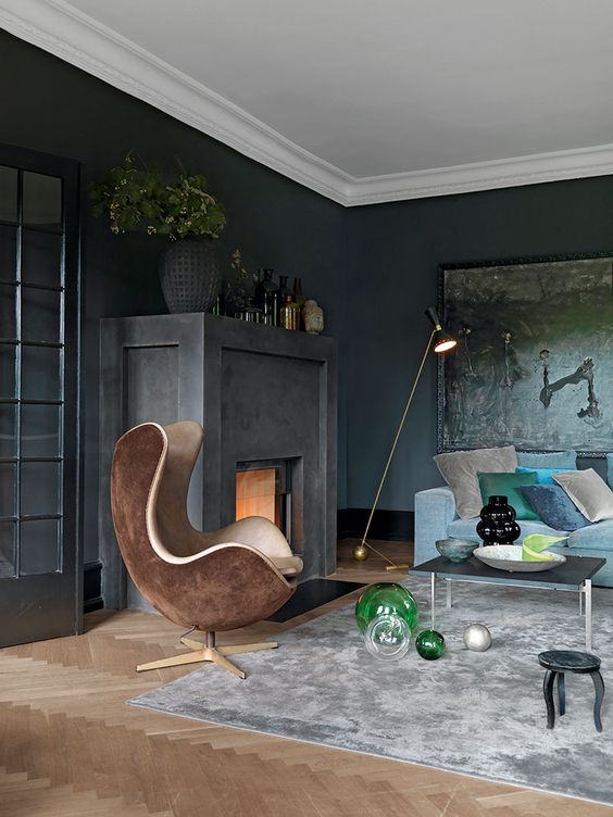 A soot living room with a firpelace, a dark artwork, a blue sofa and colorful pillows, a brown egg shaped chair and some vases
