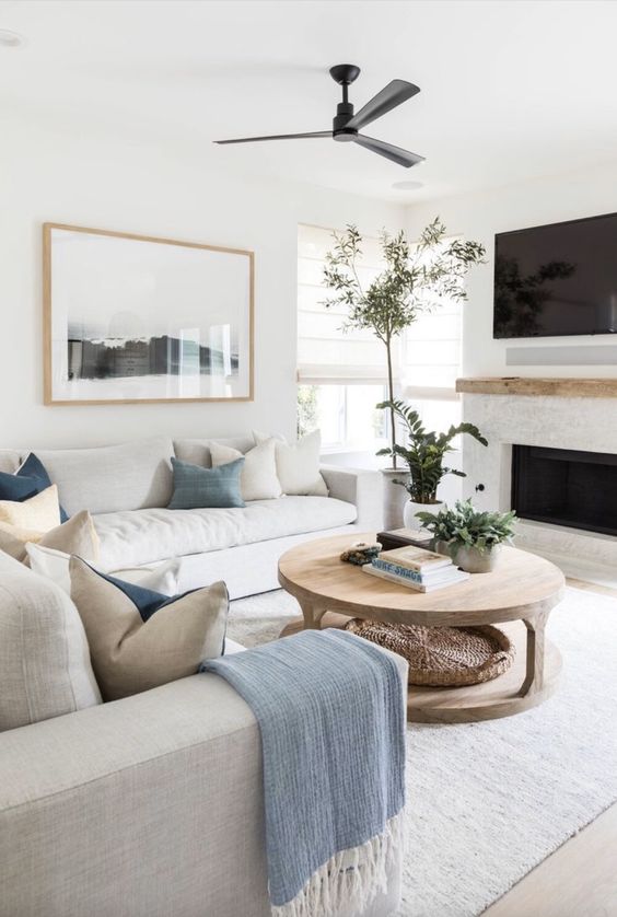 A soothing living room with a built in fireplace, neutral sofas, blue and white pillows, a wooden coffee table and greenery