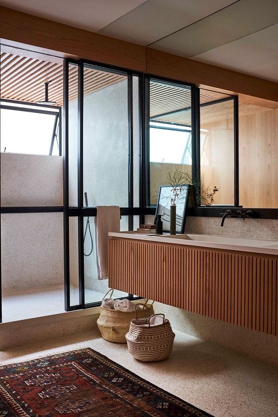 a stained fluted floating vanity with a stone countertop, a terrazzo shower space with a window, a large mirror and baskets