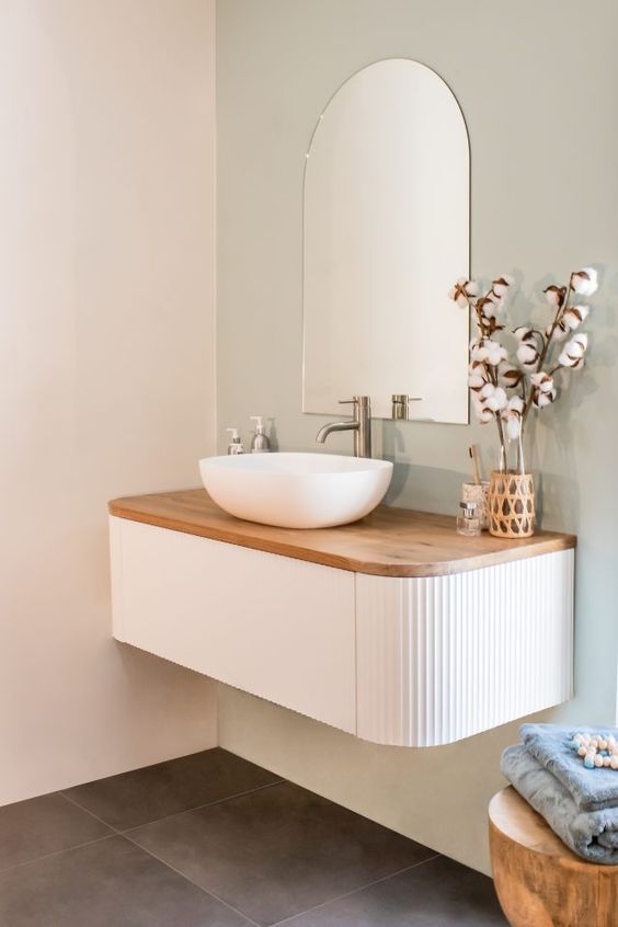 a stylish and trendy bathroom nook with an arched mirror, a white curved fluted vanity with a wooden countertop, a bowl sink