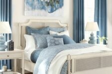a stylish blue and white bedroom with a cane bed and white nightstands, blue bedding, a printed rug, blue curtains and an artwork